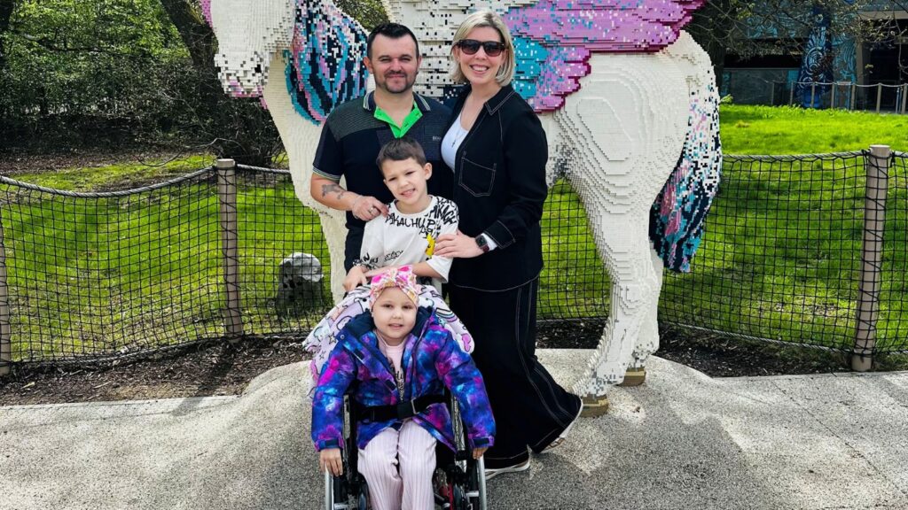Four people pose together outdoors in front of a large, colourful sculpture made of LEGO-style bricks shaped like a unicorn. A man in a dark shirt with a green collar detail and a woman wearing black clothes and sunglasses are standing at the back. In front of them are two children: one child stands slightly forward wearing a light top with printed text, while another child sits in a wheelchair wearing a bright, multicoloured jacket and a headband. The group is positioned on a paved area, with a low fence, grass, and trees visible in the background.