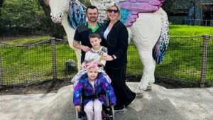 Four people pose together outdoors in front of a large, colourful sculpture made of LEGO-style bricks shaped like a unicorn. A man in a dark shirt with a green collar detail and a woman wearing black clothes and sunglasses are standing at the back. In front of them are two children: one child stands slightly forward wearing a light top with printed text, while another child sits in a wheelchair wearing a bright, multicoloured jacket and a headband. The group is positioned on a paved area, with a low fence, grass, and trees visible in the background.