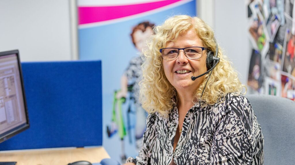 A Newlife Nurse wearing glasses and a headset sits at a desk in an office cubicle, facing the camera. A computer monitor, blue partition and colorful posters and photos are visible in the background.