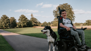 This picture shows a teenage boy in a powered wheelchair provided by Newlife with his dog in a park setting