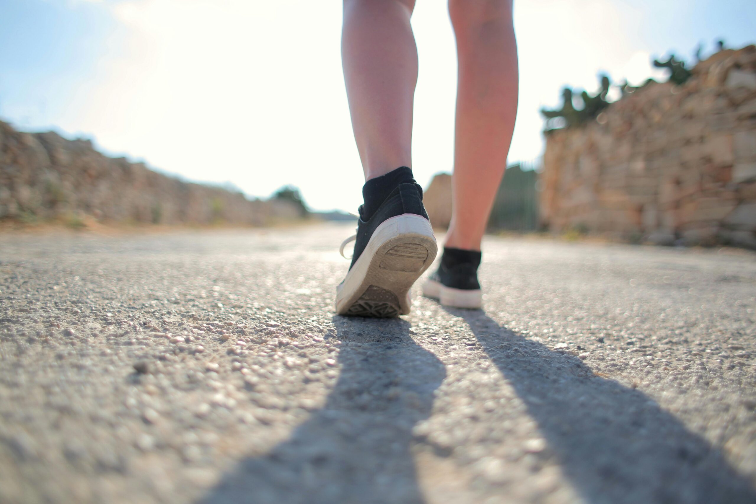 A person walks on a sunny rural road wearing trainers, casting a long shadow