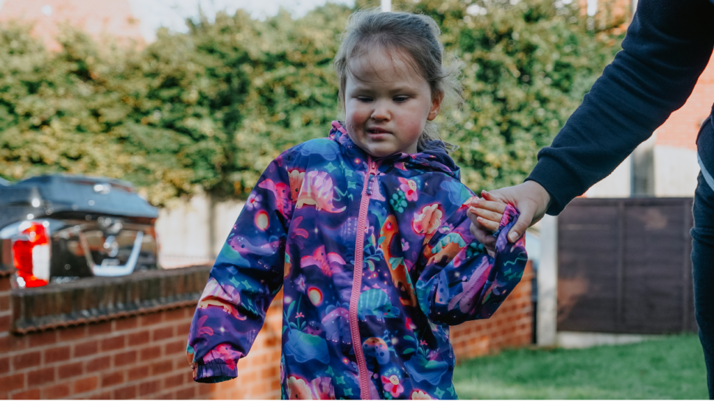 This picture shows a girl walking in a bright puddle suit. Holding hands with an adult who is guiding her.