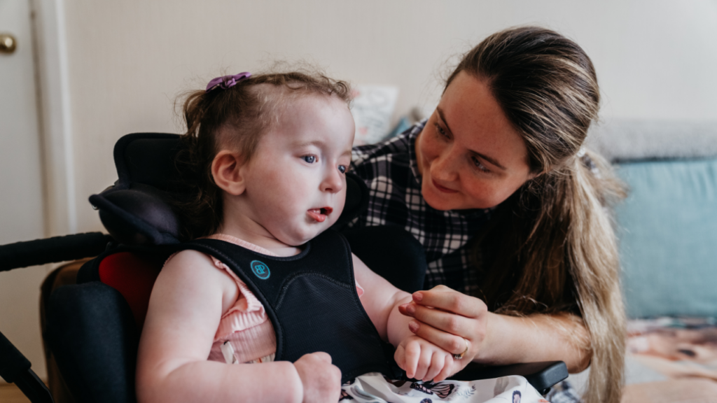 This image shows a young child in a wheelchair with her mum holding her hand and leaning in towards her.
